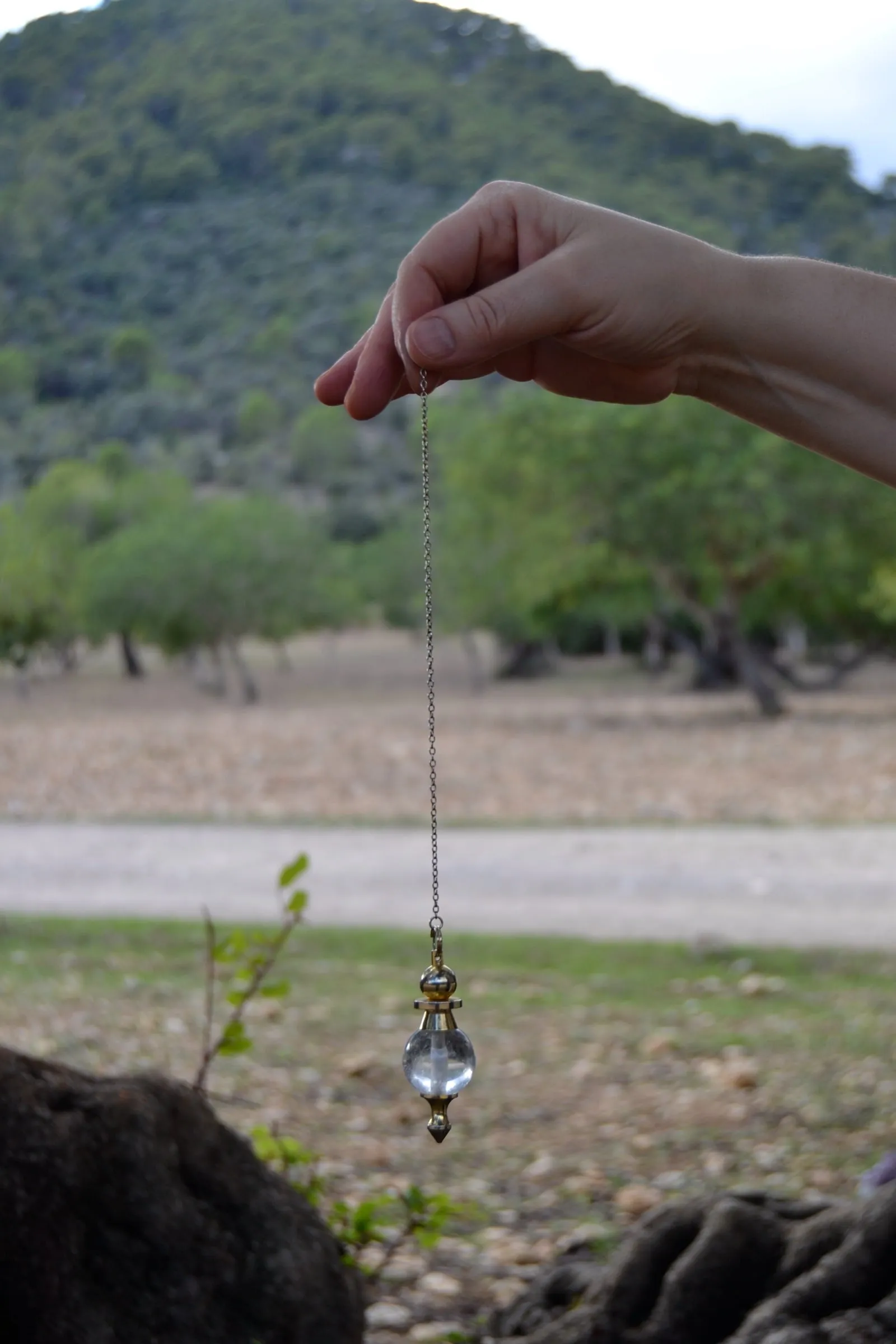 Una mano sostiene un péndulo de cristal en el campo, con un olivar detrás